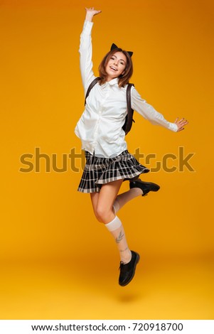 Full length portrait of a young teenage schoolgirl in uniform with backpack posing while jumping and looking at camera isolated over orange background