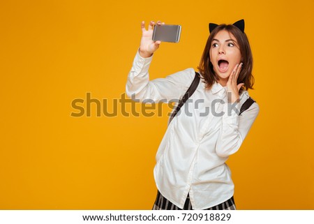 Surprised teenage schoolgirl in uniform with backpack taking a selfie while standing isolated over orange background