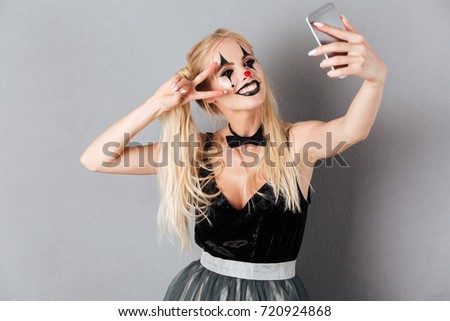 Portrait of a happy cheerful blonde woman in halloween clown make-up taking a selfie with mobile phone isolated over gray background