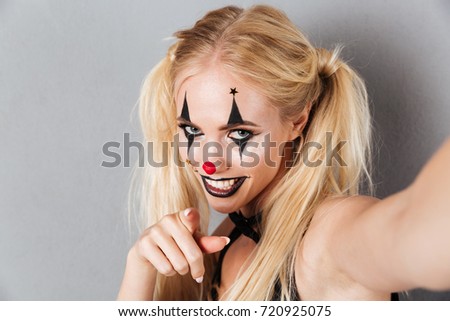 Close up portrait of a smiling blonde woman in bright halloween clown make-up taking a selfie and pointing finger at camera isolated over gray background
