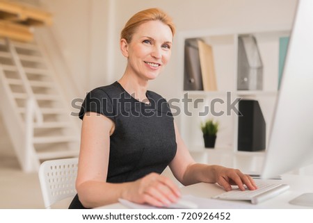 Attractive blond-haired entrepreneur posing for photography with toothy smile while checking business emails on computer, interior of modern open plan office on background