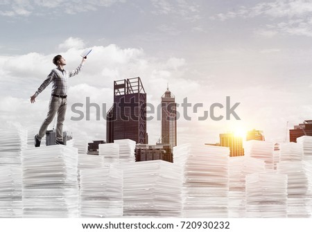Side view of man in casual wear keeping hand with book up while standing on pile of documents with cityscape and sunlight on background. Mixed media.