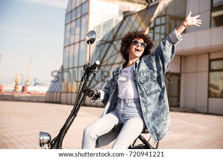 Side view of carefree curly girl in sunglasses sitting on modern motorbike outdoors and waving away