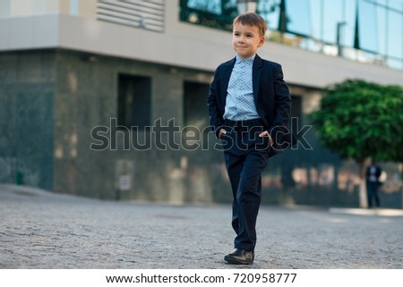 Concept future businessman. Smiling boy in classic modern dark blue business costume and baby blue shirt walking on pedestrian street near building and trees with hands in pockets.