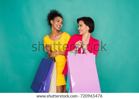 Happy multiethnic girlfriends with shopping bags. Two excited shopaholics at turquoise sudio background with copy space