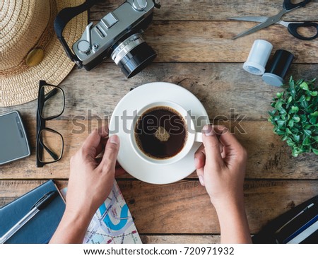 Travel concept with hands holding a coffee cup, retro camera, smartphone, notebook, pen, map, straw hat on wooden background.