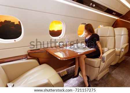 young beautiful woman in Luxury interior in bright colors of genuine leather in the business jet, sky and rays of sunset through the porthole Royalty-Free Stock Photo #721157275