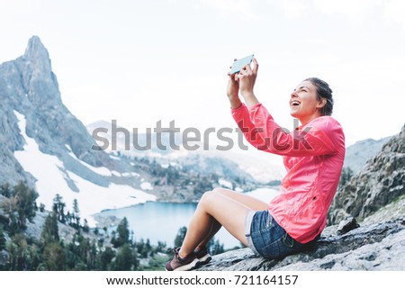 Excited woman sitting high in mountains and taking photo using her mobile phone. Risky rock climbing in peaceful wilderness area. Enjoying amazing snowy lake view