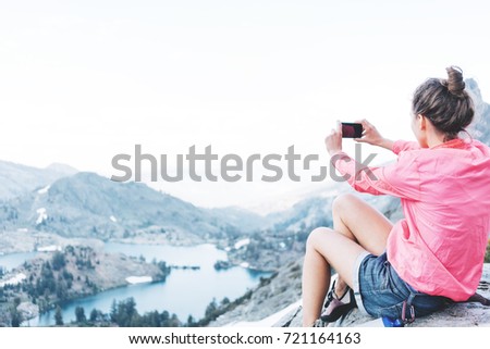 Brave woman sitting high in mountains and taking photo using her phone. Risky rock climbing in peaceful wilderness area. Enjoying amazing snowy lake view