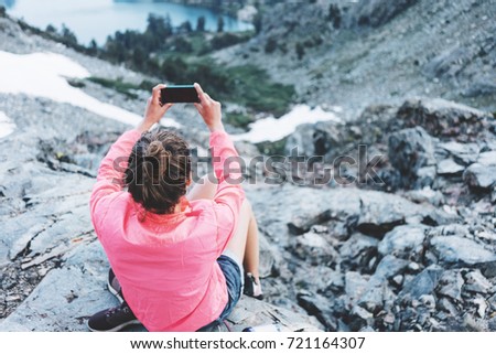 Brave woman sitting high in mountains and taking photo using her smartphone. Risky rock climbing in peaceful wilderness area. Enjoying amazing snowy lake view