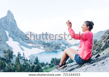 Young brave woman sitting high in mountains and taking photo using her phone. Risky rock climbing in peaceful wilderness area. Enjoying amazing snowy lake view