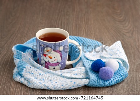 Tea in the winter. A picture of a snowman on a mug. Tea and a knitted scarf on a cold winter day. New Year and Winter Composition. Tea and a scarf create coziness in the winter.