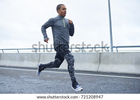 Freeze action shot of confident young black athletic man wearing trendy running shoes and sports clothes exercising outdoors. Picture of dark-skinned sportsman running on autumn day at stadium