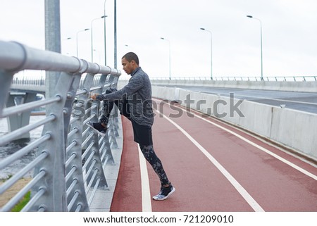 Profile shot of serious young Afro American male runner in stylish sports clothes and running shoes stretching legs at stadium, warming up muscles before run. Fitness, sports and self-determination