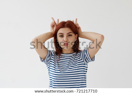 Waist up picture of cute playful funny teenage girl with red colored hair pouting and holding hands behind her head like bunny bears or horns, having fun. People, youth and lifestyle concept