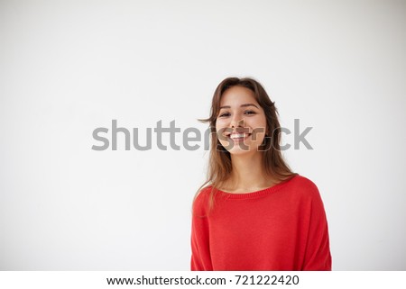Picture of attractive cheerful young Hispanic lady dressed in oversize red sweater grinning broadly at camera, her eyes expressing joy. Positive friendly looking girl posing isolated in studio