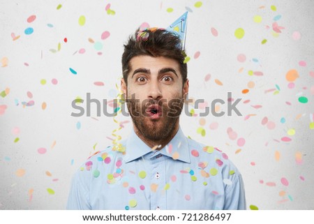 People, joy, birthday, anniversary, fun and party concept. Picture of handsome astonished young man with cone hat on his head celebrating birthday among friends, amazed with unexpected gift