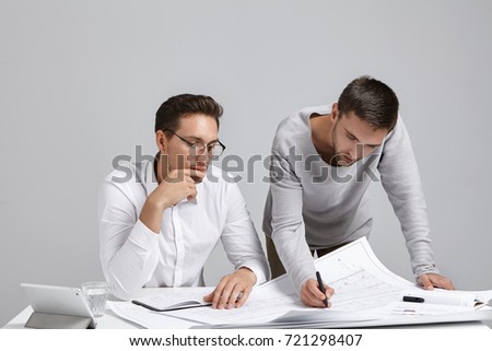 Portrait of two creative arhitects sit at workplace, discuss house project, look attentively at blueprint, make corrections, decide how to do better, use modern tablet for surfing construction site