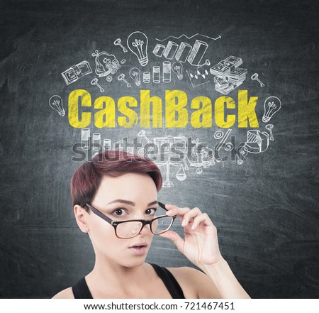 Portrait of a young surprised woman with short red hair taking off her glasses. She is standing near a blackboard with a white and yellow cashback sketch drawn on it.