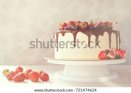 Cake in chocolate, decorated with slices strawberries, blueberries and figs on a white wooden table in the morning sun. Picture for a menu or a confectionery catalog with copy space. Soft light.