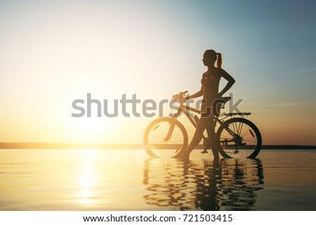 The strong blonde woman in a colorful suit stands near the bicycle in the water at sunset on a warm summer day. Fitness concept. Sky background