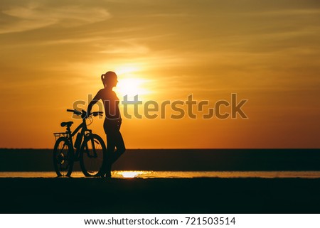The silhouette of a sporty girl in a suit standing near a bicycle in the water at sunset on a warm summer day. Fitness concept. Sky background