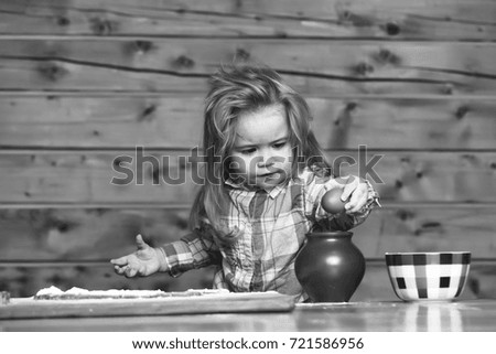 adorable small child chef or cute happy baby boy with blonde hair in fashionable chekered shirt cooking on board flour and dough near bowl and egg for cookies on wooden background