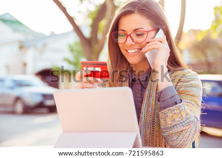Smiling woman sitting outdoors talking on mobile phone making online payment on her tablet computer outside on a sunny autumn day  Royalty-Free Stock Photo #721595863
