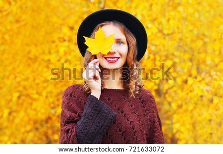 Autumn portrait of happy smiling woman covering her eyes with yellow maple leaves wearing round hat in the park 