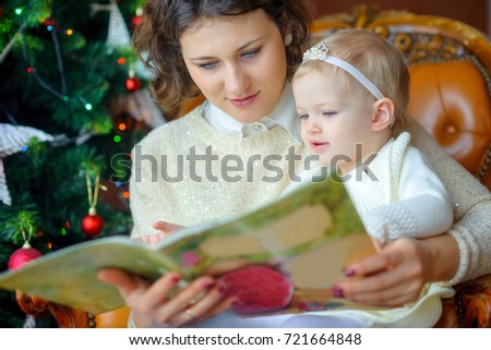 Mum reads fairy tales to a small daughter sitting near a festive Christmas tree
