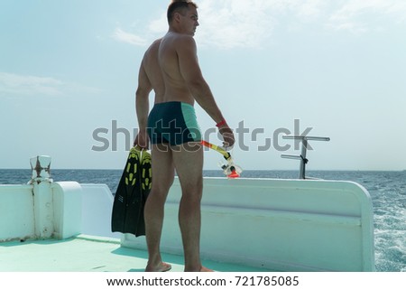 The guy stands in the slats in the stern of the yacht with fins and a mask in his hands. The guy is preparing to dive under water.