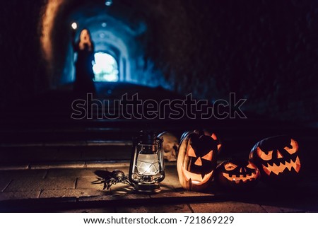 Happy Halloween! Scary flaming pumpkins with horrible faces in dark cave. Lamp and skull lying nearby. Black witch with torch in hand is on the background.