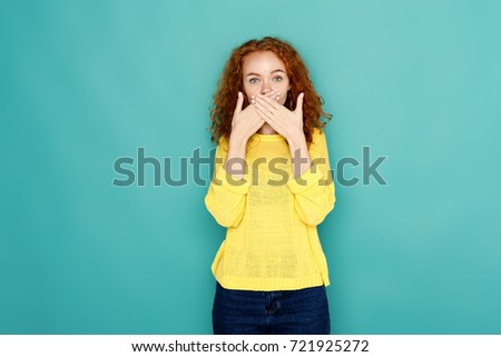 Keep silence. Redhead woman covering mouth with hands while posing to camera on blue studio background. Girl close lips with palms, speak no evil concept