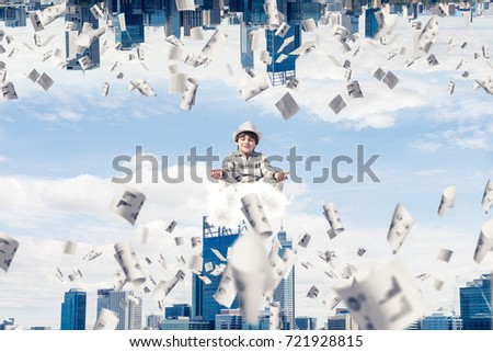 Young boy keeping eyes closed and looking concentrated while meditating on cloud among flying papers and between two urban worlds.