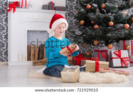 Cute happy boy in santa hat unwrap christmas present box on holiday morning in beautiful room interior. Male child open Xmas gifts near big decorated fir tree and fireplace. Winter holidays concept