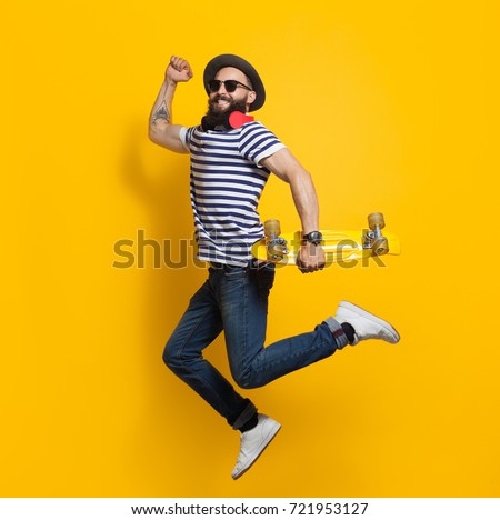 Side view of stylish hipster man with cruiser board jumping on colorful yellow background.