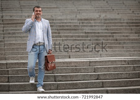 Successful and professional businessman in a light suit, walking with a brown briefcase. A mature business employee texting with a new phone. Technological progress concept. Copy space.