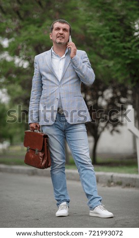 A professional executive businessman in comfortable clothes walking down the street on the blurred urban background. Office worker with a briefcase talking on a phone. Financial occupation concept.