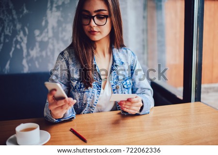 Serious young woman in stylish eyeglasses holding business card in hand and dialing mobile number on modern smartphone.Successful female typing phone information on cellular sitting at table in cafe