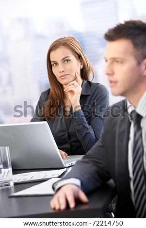 Portrait of young businesswoman sitting at table, using laptop, man in background.?