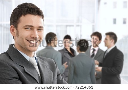 Smiling confident businessman portrait, group of businesspeople chatting in background.?