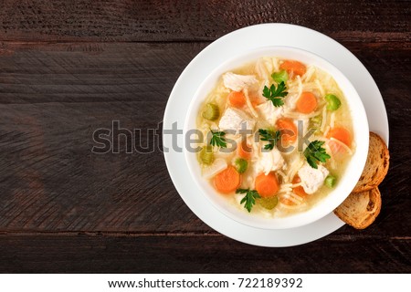 An overhead photo of a plate of chicken, vegetables, and noodles soup, shot from above on a dark rustic texture with slices of bread, and a place for text