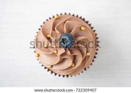 Chocolate cupcake with whipped chocolate cream, decorated fresh blueberry, gold confectionery sprinkling on white wooden table. Picture for a menu or a confectionery catalog. Top view.