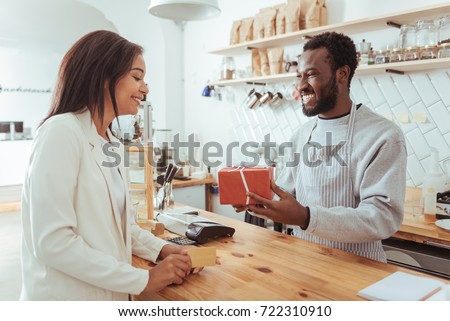 Best service. Joyful pleasant barista giving his pretty female customer a box with her order while the woman holding a credit card, being ready to pay