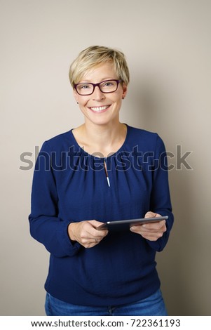 Friendly blond woman wearing glasses with a beaming smile standing holding a tablet over a neutral studio background