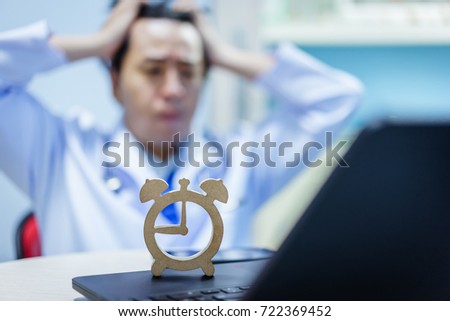 Close up wooden clock model put on the notebook computer and blur asian doctor sitting and hold the head with both hand, stress, headache, worried, strain.