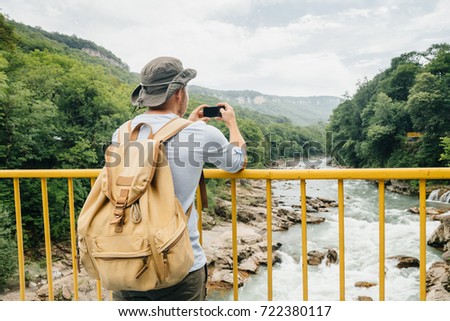 young traveler in a hat with a backpack full back stopped to take a photo of a mountain river. concept of travel and photography.
