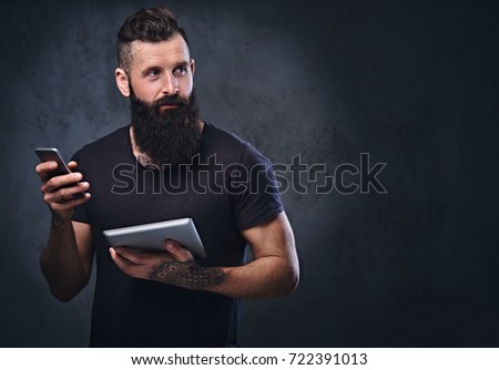 The tattooed, bearded hipster male holds tablet PC and smartphone on grey background.