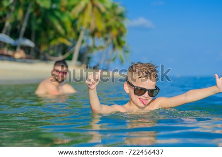 Close-up portrait in tropical water: seven years old cutest blond boy lying on the water surface, held up thumbs and show tongue. He is in sunglasses, his hair is wet. On background sits his father