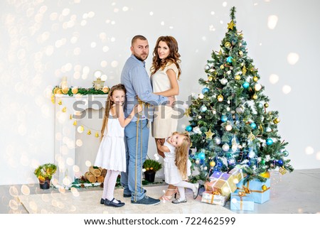 Happy family, parents hugging with their daughters by the white fireplace with an elegant Christmas tree in garlands of lights. A young father and a beautiful mother are holding hands, little sisters 